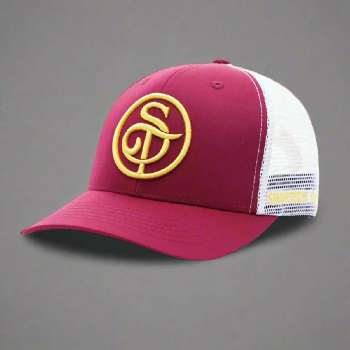 The Smartech Outfit Burgundy Trucker Mesh Cap is a premium cap featuring a burgundy and white design with a yellow embroidered logo of intertwined letters on the front. It offers a mesh back for ventilation, an adjustable snapback closure, and UV light protection for extra durability.